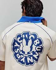 Cream jersey polo with royal blue collar, contrast shoulder piping, and a large back graphic featuring the Psycho Bunny skull logo and text, plus a small embroidered collar logo. Mens Toby Jersey. (3)
