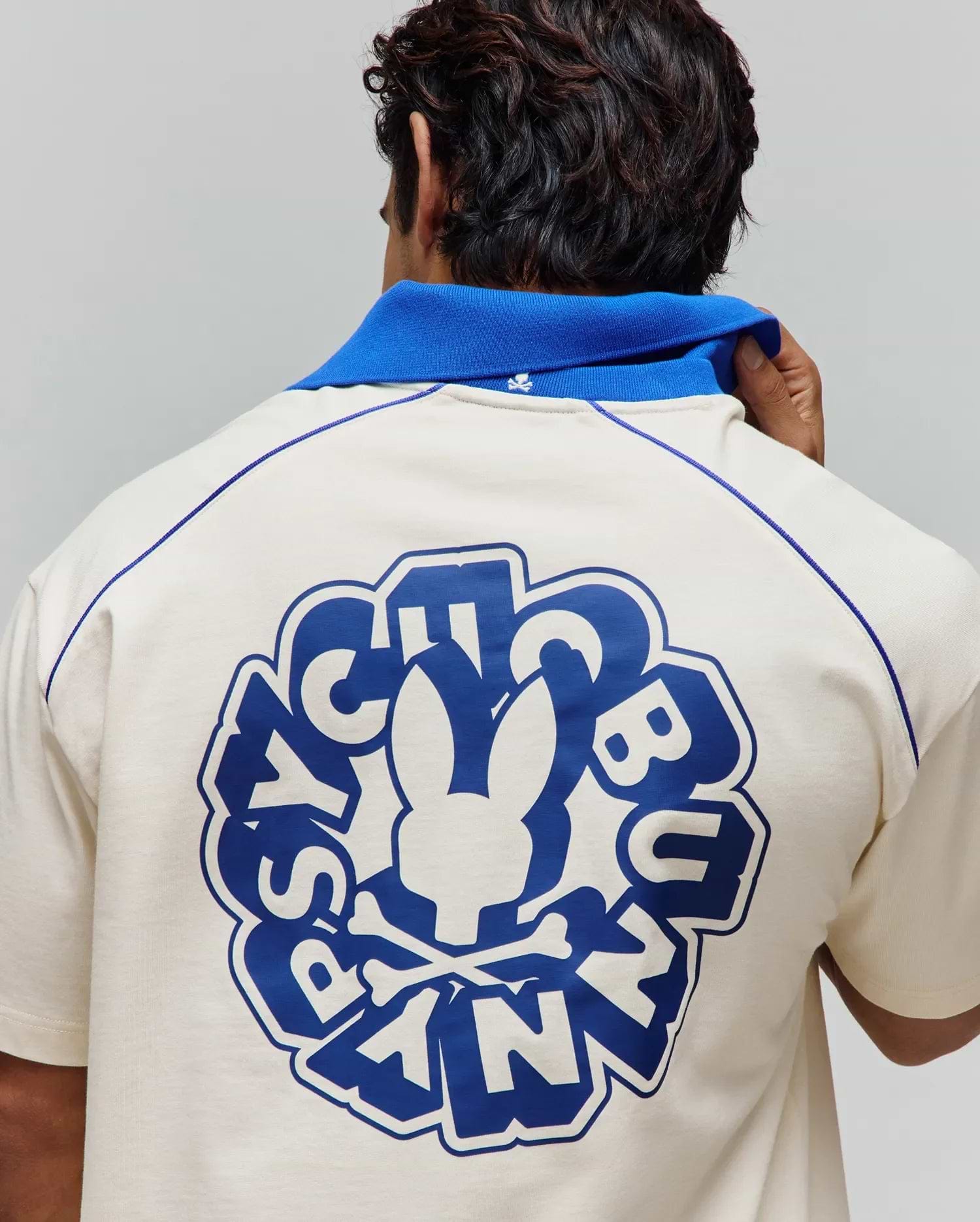 Cream jersey polo with royal blue collar, contrast shoulder piping, and a large back graphic featuring the Psycho Bunny skull logo and text, plus a small embroidered collar logo. Mens Toby Jersey. (3)