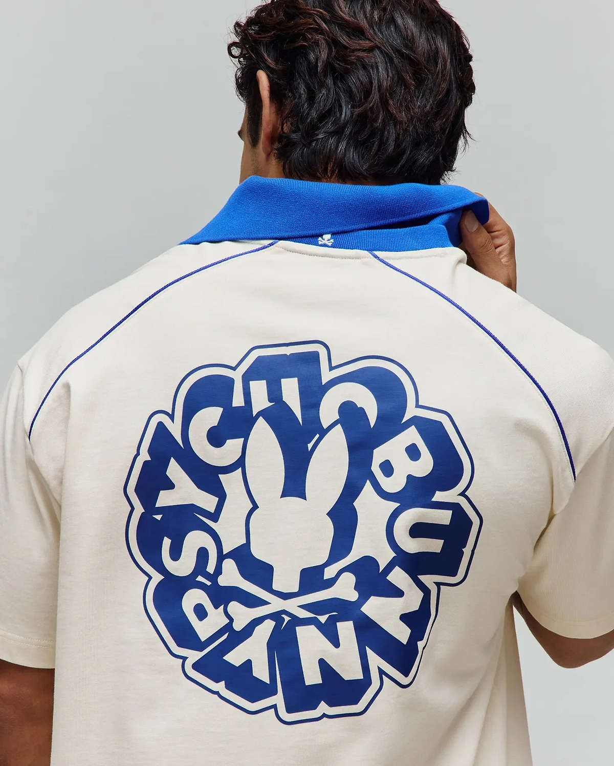 Cream jersey polo with royal blue collar, contrast shoulder piping, and a large back graphic featuring the Psycho Bunny skull logo and text, plus a small embroidered collar logo. Mens Toby Jersey. (3)