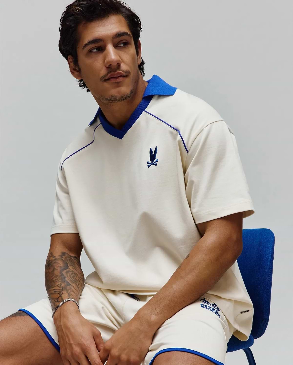 Mens Toby Jersey: Relaxed cream jersey polo with royal blue spread collar, V-neck, piping, and small embroidered bunny skull on chest; matching shorts have royal blue trim and graphic logo. (4)