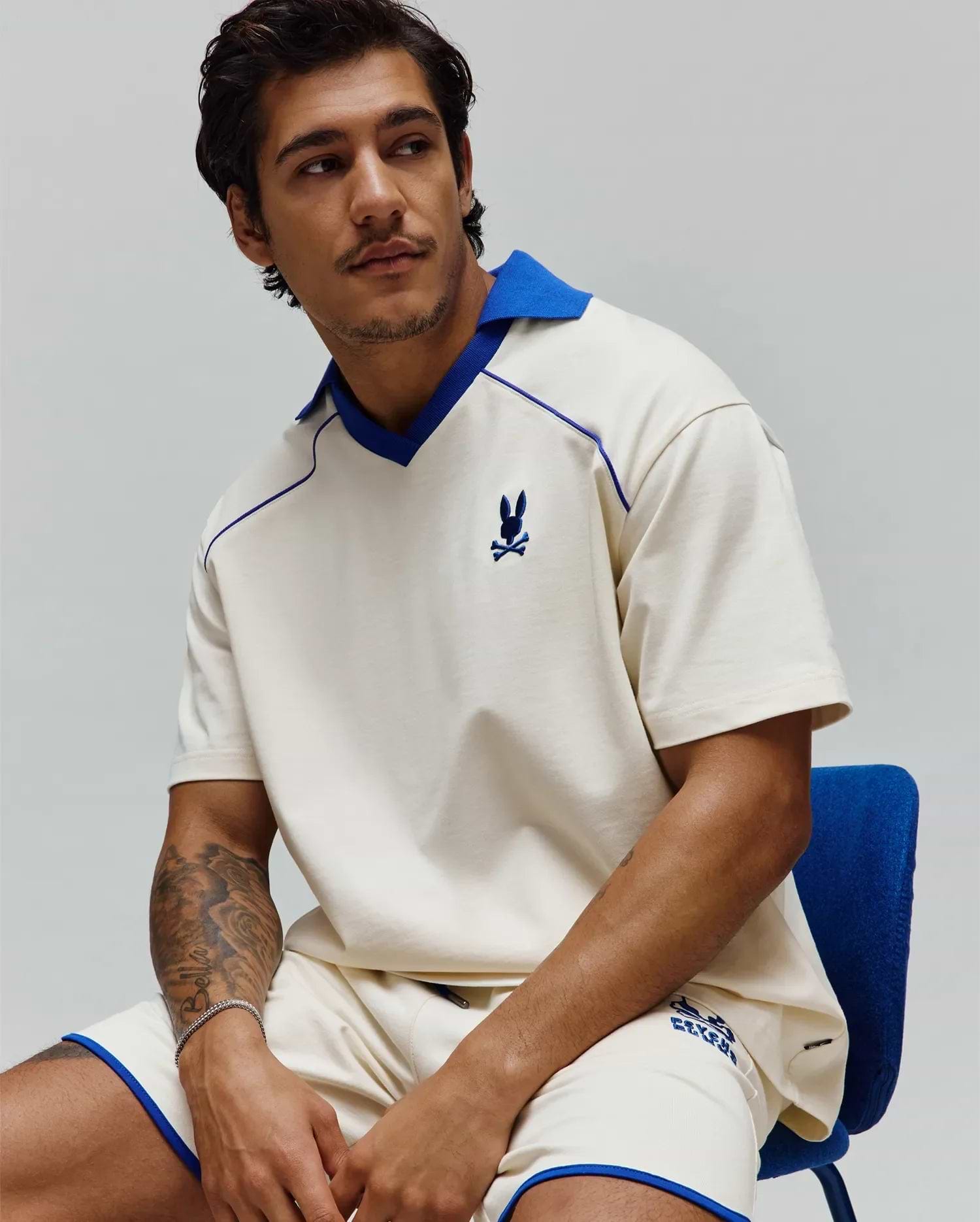 Mens Toby Jersey: Relaxed cream jersey polo with royal blue spread collar, V-neck, piping, and small embroidered bunny skull on chest; matching shorts have royal blue trim and graphic logo. (4)