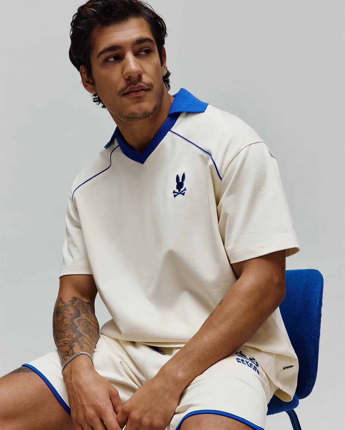 Mens Toby Jersey: Relaxed cream jersey polo with royal blue spread collar, V-neck, piping, and small embroidered bunny skull on chest; matching shorts have royal blue trim and graphic logo. (4)