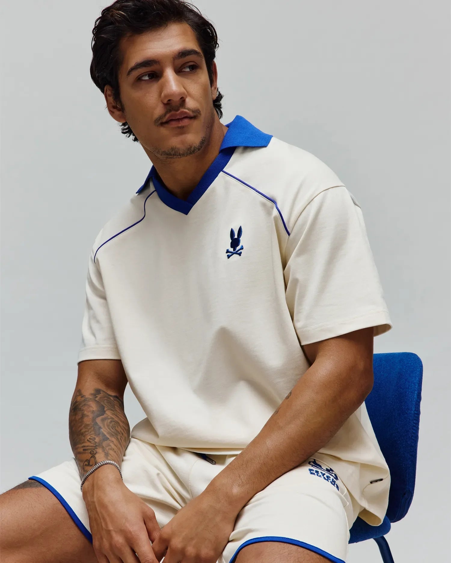 Mens Toby Jersey: Relaxed cream jersey polo with royal blue spread collar, V-neck, piping, and small embroidered bunny skull on chest; matching shorts have royal blue trim and graphic logo. (4)