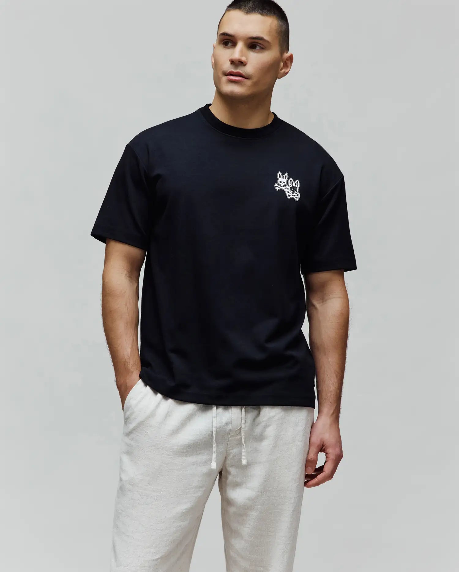 Black Mens Luke Back Graphic Tee, a relaxed fit jersey t-shirt with a small white embroidered Psycho Bunny double-bunny skull logo on the left chest and a graphic on the back.