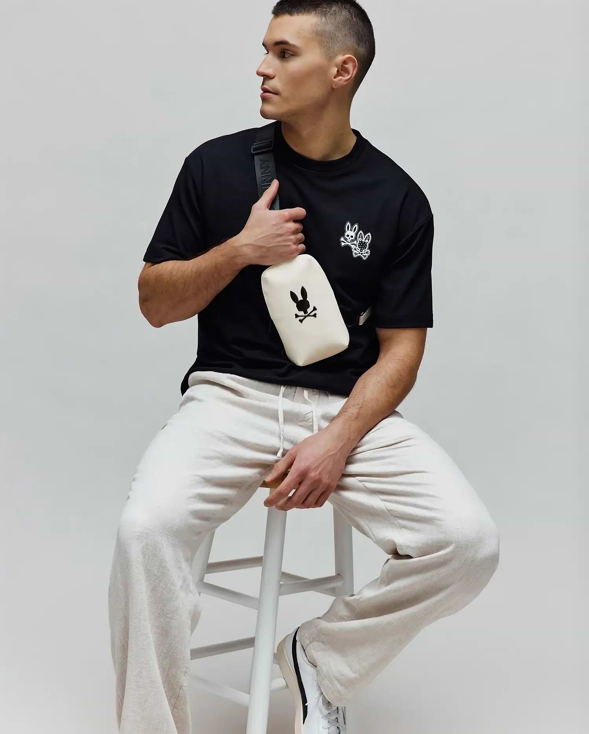 Black jersey Mens Luke Back Graphic Tee, classic fit, with white embroidered Psycho Bunny skull and bow tie logos on the chest, paired with relaxed light beige drawstring joggers.