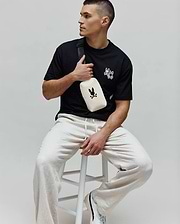 Black jersey Mens Luke Back Graphic Tee, classic fit, with white embroidered Psycho Bunny skull and bow tie logos on the chest, paired with relaxed light beige drawstring joggers.