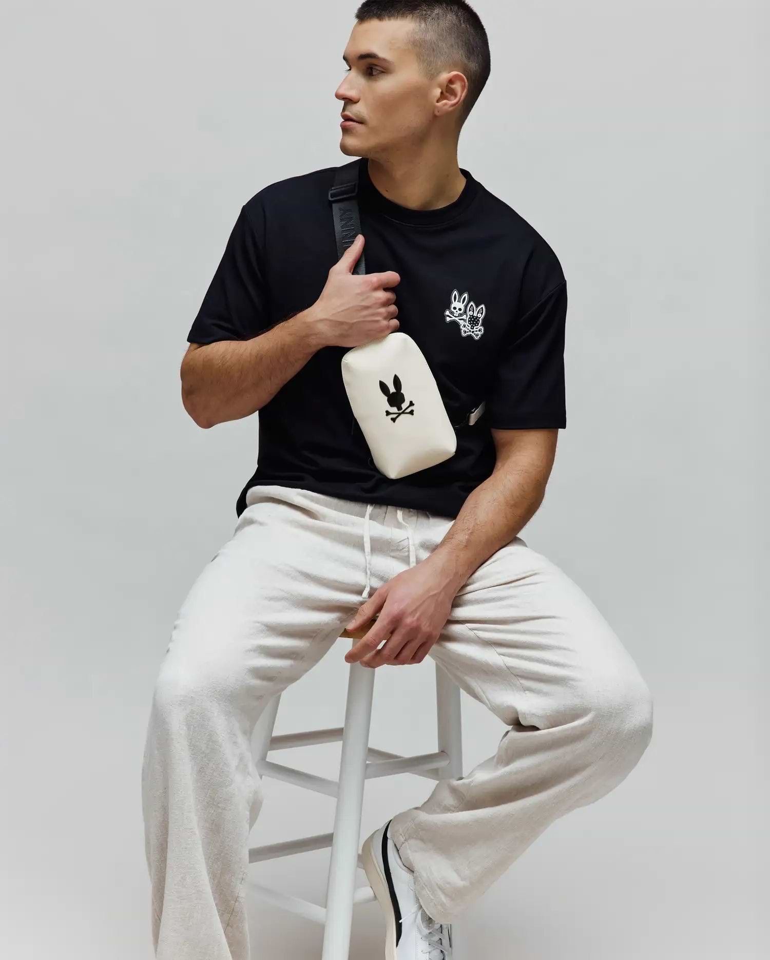 Black jersey Mens Luke Back Graphic Tee, classic fit, with white embroidered Psycho Bunny skull and bow tie logos on the chest, paired with relaxed light beige drawstring joggers.