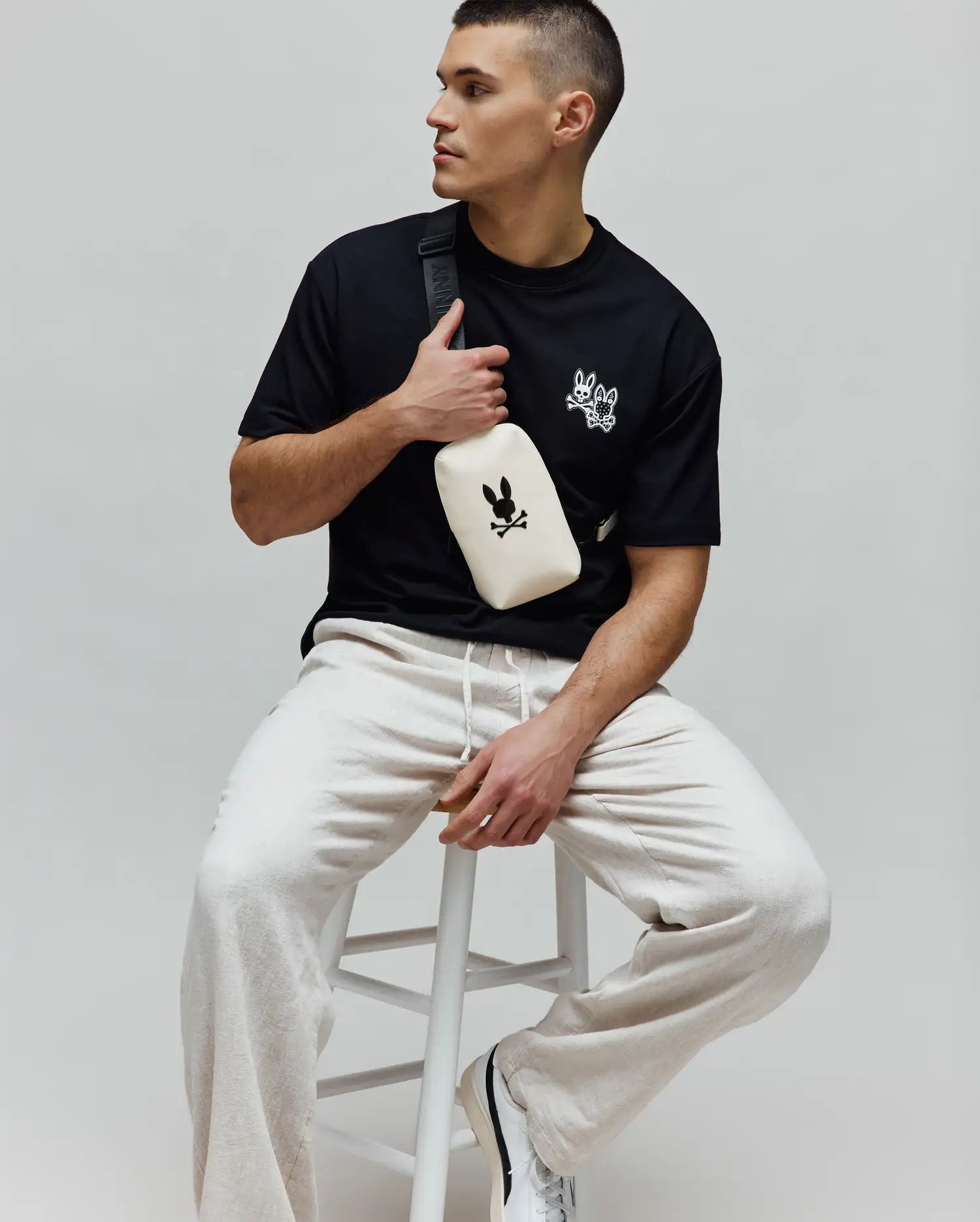 Black jersey Mens Luke Back Graphic Tee, classic fit, with white embroidered Psycho Bunny skull and bow tie logos on the chest, paired with relaxed light beige drawstring joggers.