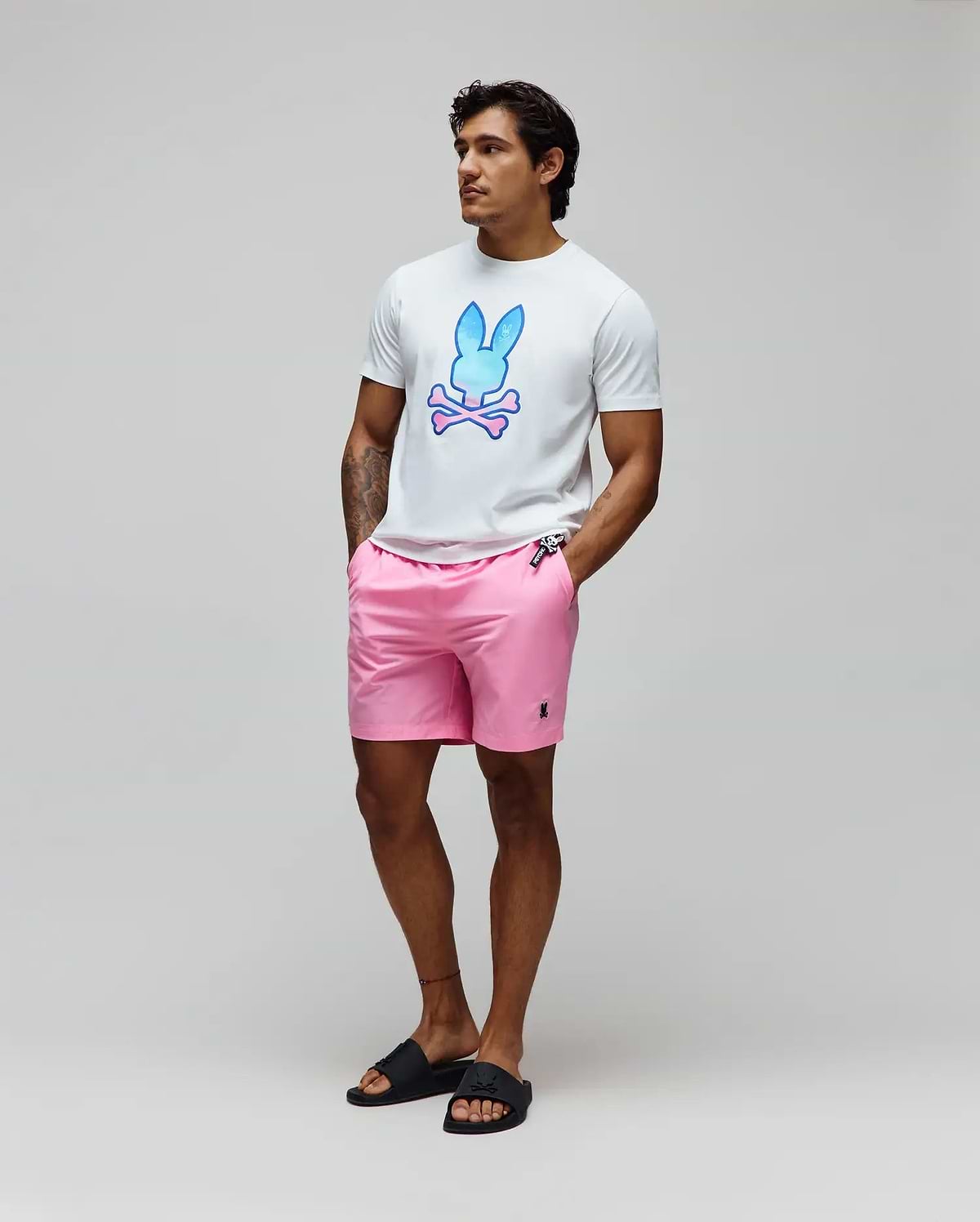 Mens Caspian Graphic Tee, a white classic fit jersey t-shirt with a large blue-pink gradient Psycho Bunny skull graphic on the chest, paired with pink woven shorts featuring a small embroidered black bunny logo.