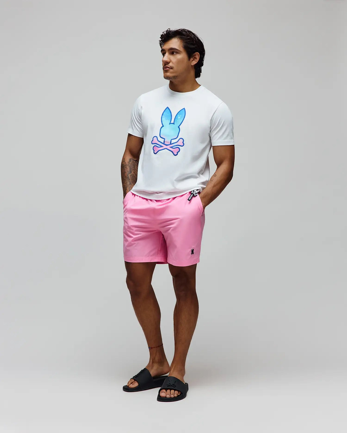 Mens Caspian Graphic Tee, a white classic fit jersey t-shirt with a large blue-pink gradient Psycho Bunny skull graphic on the chest, paired with pink woven shorts featuring a small embroidered black bunny logo.