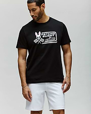 A man stands against a light gray background, looking down and to the side, wearing the Psycho Bunny MENS RACING GRAPHIC TEE (MT0200243) in black with a pink and white Bunny graphic, paired with white athletic shorts. (2)