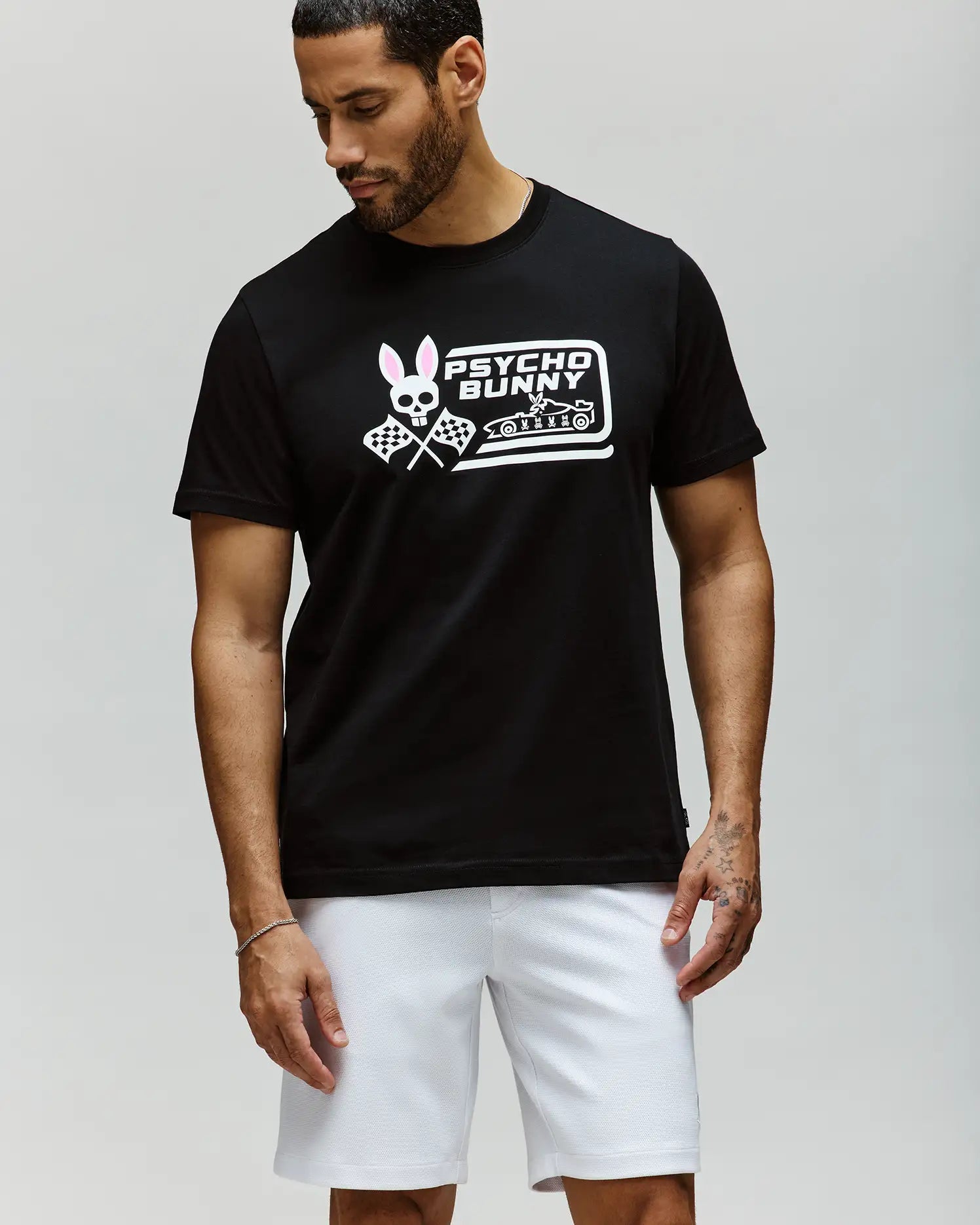 A man stands against a light gray background, looking down and to the side, wearing the Psycho Bunny MENS RACING GRAPHIC TEE (MT0200243) in black with a pink and white Bunny graphic, paired with white athletic shorts. (2)