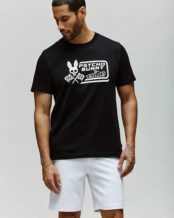 A man stands against a light gray background, looking down and to the side, wearing the Psycho Bunny MENS RACING GRAPHIC TEE (MT0200243) in black with a pink and white Bunny graphic, paired with white athletic shorts. (2)