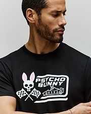 A bearded man looks to the side, wearing Psycho Bunny’s MENS RACING GRAPHIC TEE (MT0200243), a black Pima cotton shirt featuring a white Bunny logo with skull and ears, crossed checkered flags, and “PSYCHO BUNNY” text. (3)