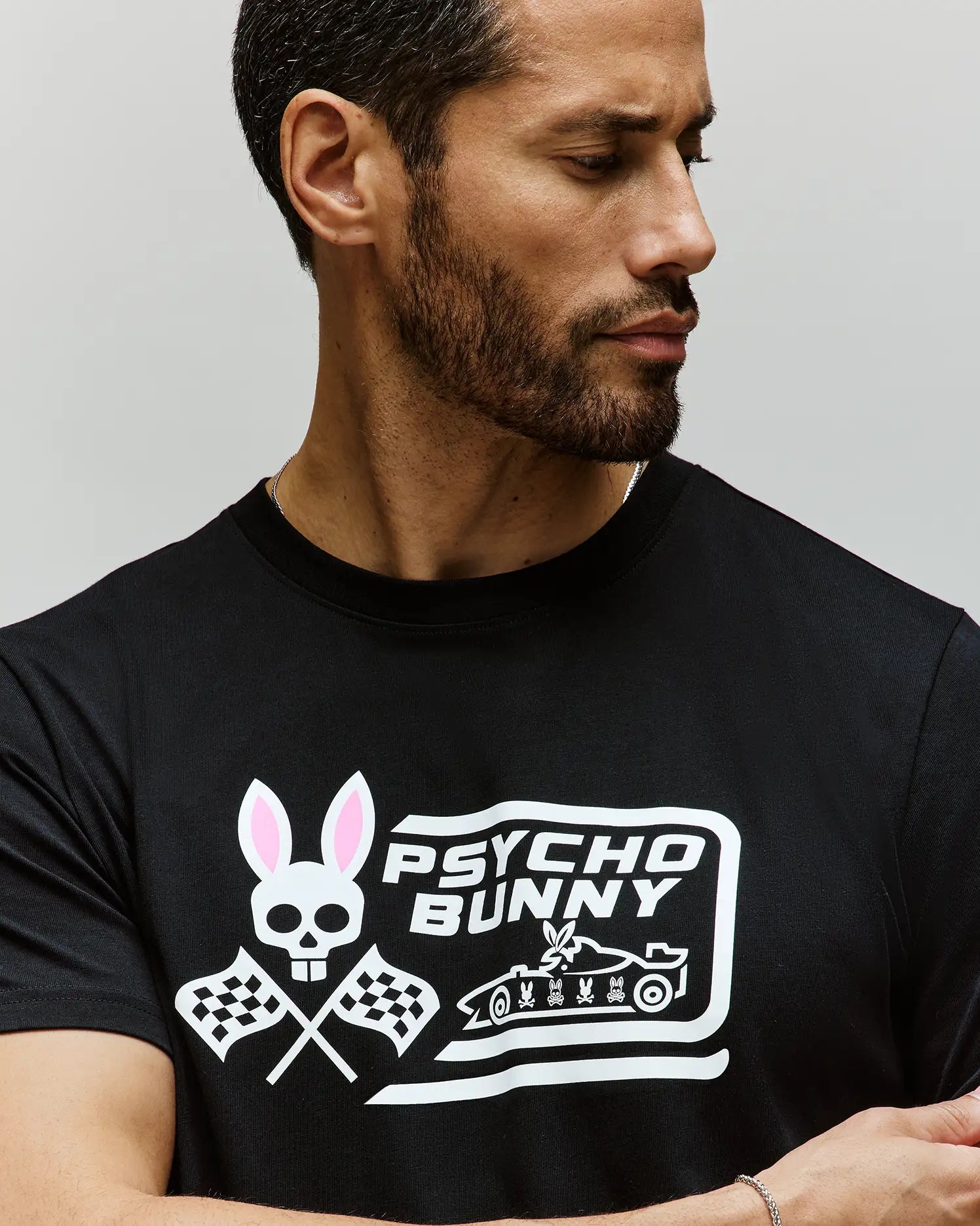 A bearded man looks to the side, wearing Psycho Bunny’s MENS RACING GRAPHIC TEE (MT0200243), a black Pima cotton shirt featuring a white Bunny logo with skull and ears, crossed checkered flags, and “PSYCHO BUNNY” text. (3)