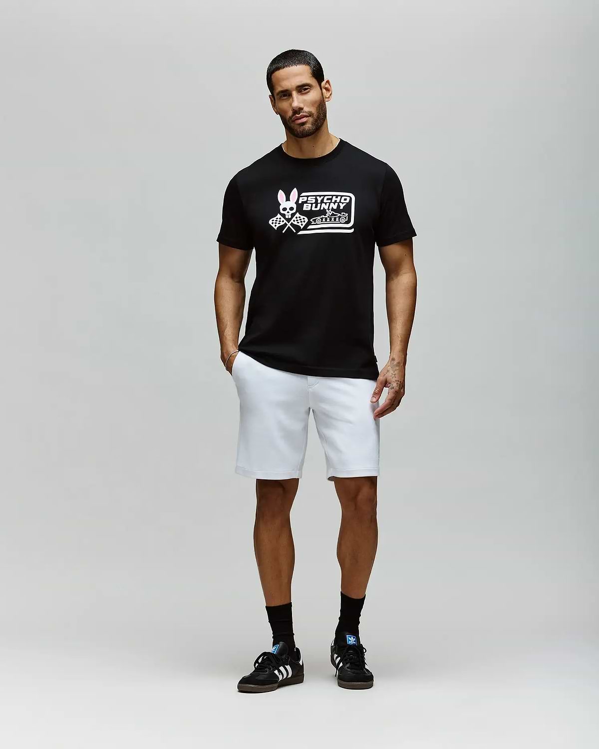 A man with short hair and a beard stands against a plain light gray background, wearing the Psycho Bunny MENS RACING GRAPHIC TEE (MT0200243), white shorts, and black sneakers with white stripes; his left hand is in his pocket. (4)
