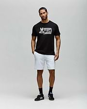 A man with short hair and a beard stands against a plain light gray background, wearing the Psycho Bunny MENS RACING GRAPHIC TEE (MT0200243), white shorts, and black sneakers with white stripes; his left hand is in his pocket. (4)