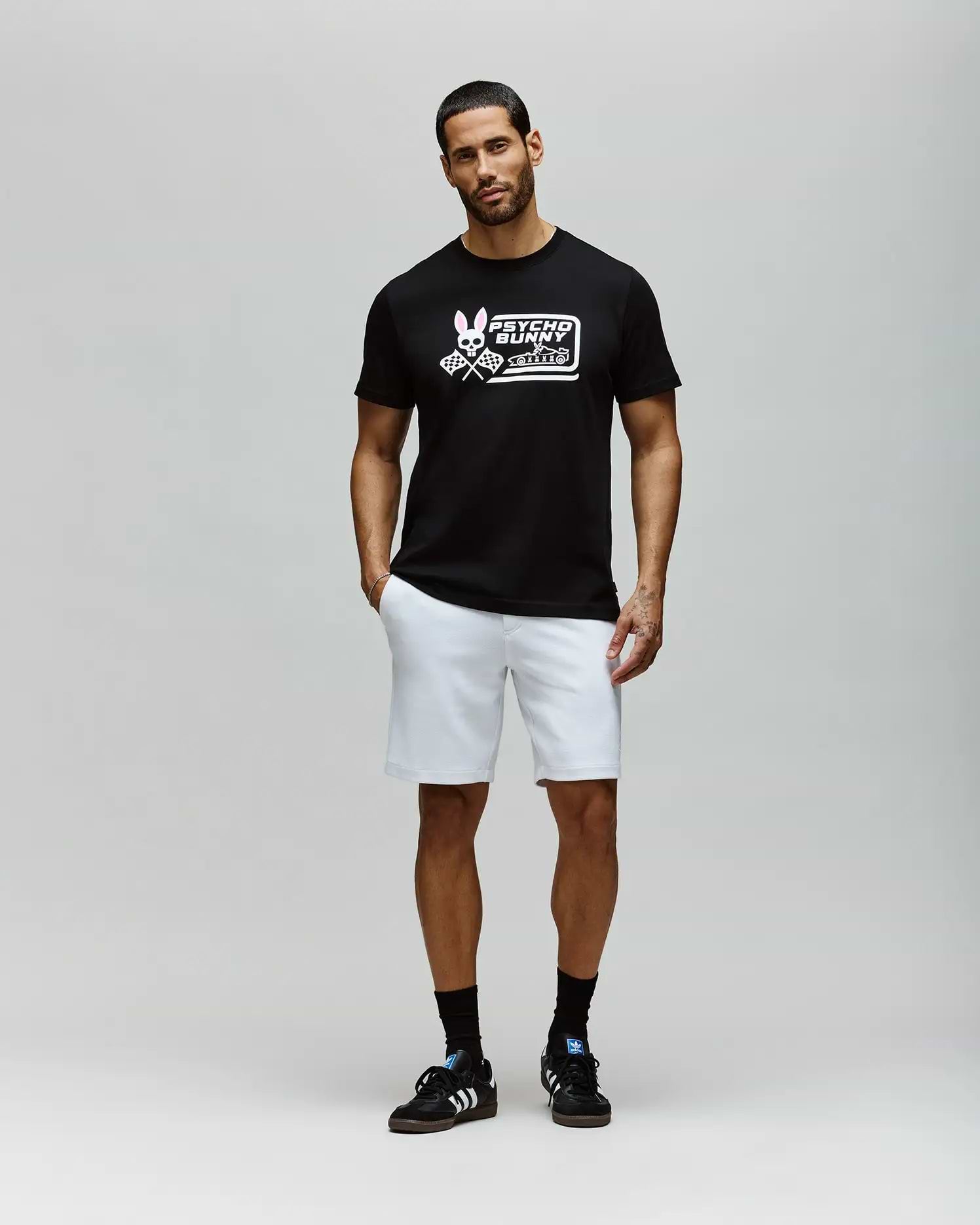 A man with short hair and a beard stands against a plain light gray background, wearing the Psycho Bunny MENS RACING GRAPHIC TEE (MT0200243), white shorts, and black sneakers with white stripes; his left hand is in his pocket. (4)