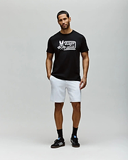 A man with short hair and a beard stands against a plain light gray background, wearing the Psycho Bunny MENS RACING GRAPHIC TEE (MT0200243), white shorts, and black sneakers with white stripes; his left hand is in his pocket. (4)