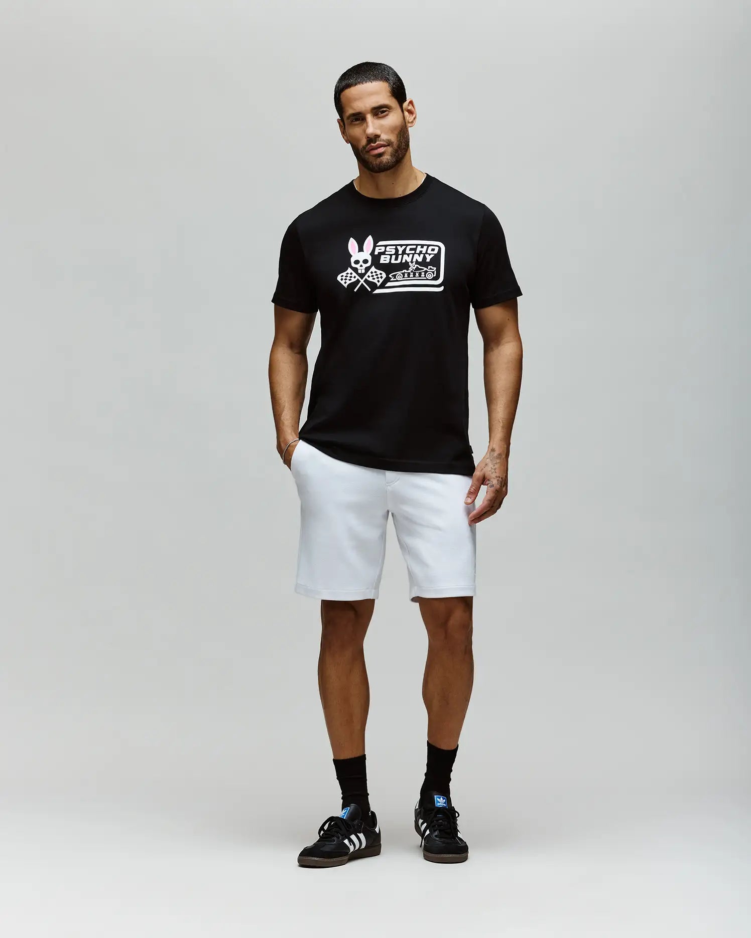 A man with short hair and a beard stands against a plain light gray background, wearing the Psycho Bunny MENS RACING GRAPHIC TEE (MT0200243), white shorts, and black sneakers with white stripes; his left hand is in his pocket. (4)
