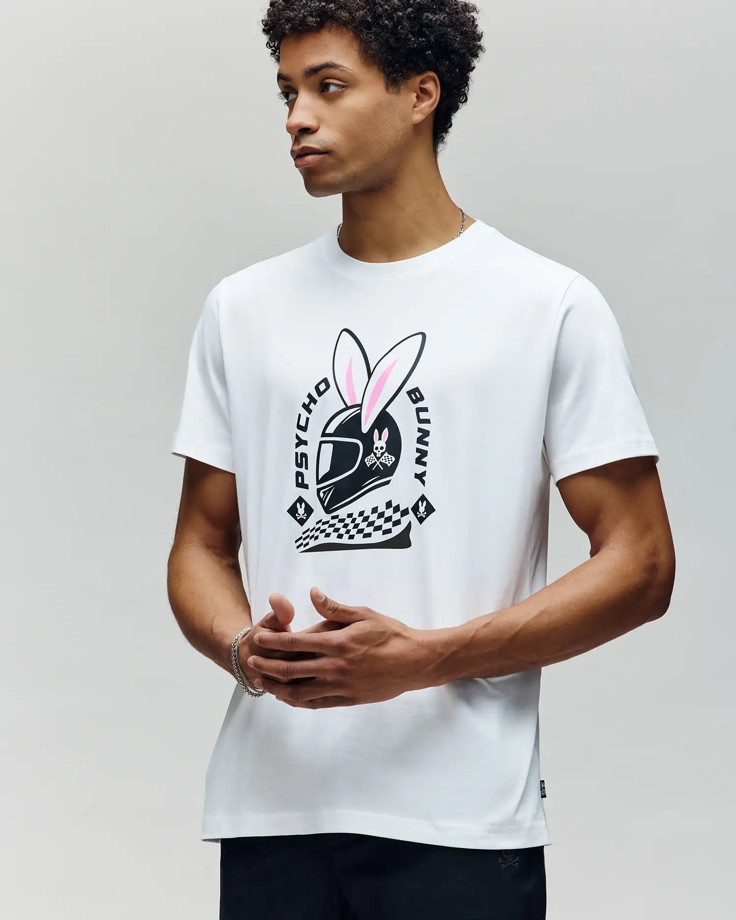 A man stands against a plain light background wearing the Psycho Bunny MENS RACING BUNNY GRAPHIC TEE (MT0200245), a race-day-ready white shirt featuring a large helmeted bunny graphic, checkered patterns, and "Psycho Bunny" text on the front. (2)