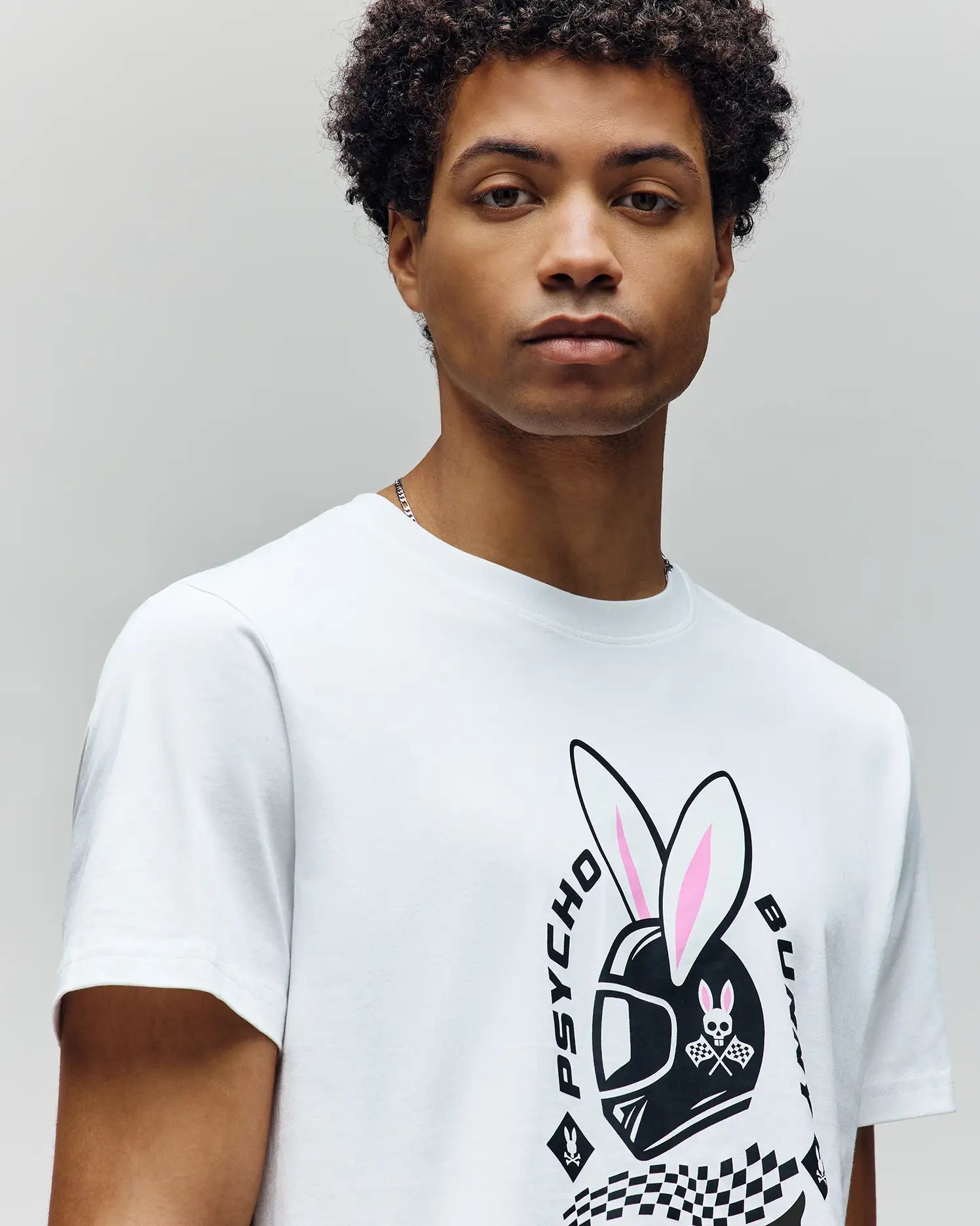 A young man with short, curly hair wears the Psycho Bunny MENS RACING BUNNY GRAPHIC TEE (MT0200245), a white, athletic-fit shirt featuring a helmet with bunny ears, "PSYCHO" and "BUNNY" text, and a checkered flag graphic. (3)
