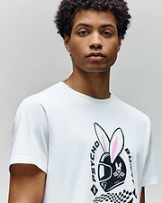 A young man with short, curly hair wears the Psycho Bunny MENS RACING BUNNY GRAPHIC TEE (MT0200245), a white, athletic-fit shirt featuring a helmet with bunny ears, "PSYCHO" and "BUNNY" text, and a checkered flag graphic. (3)