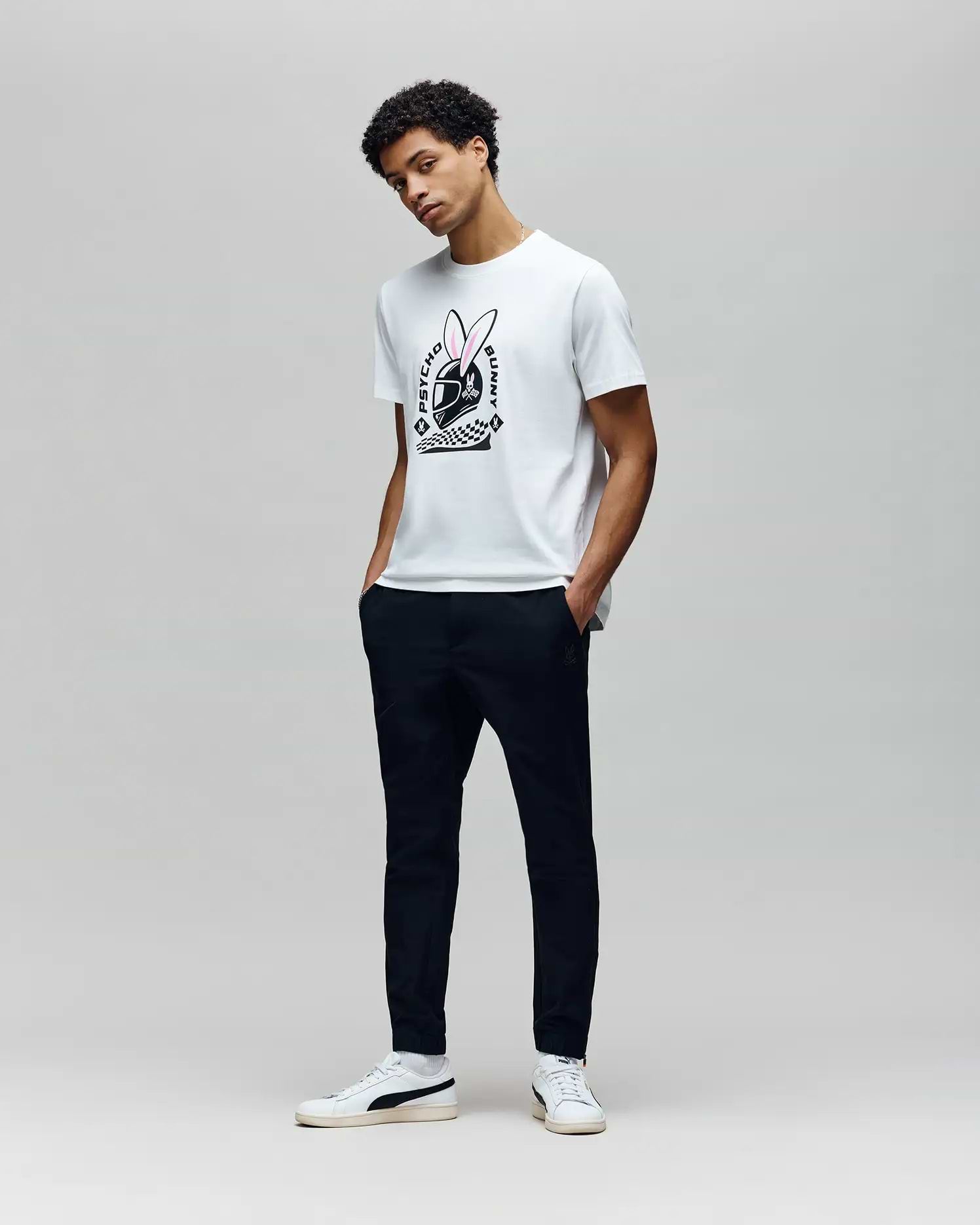 A man poses against a light gray background wearing the Psycho Bunny MENS RACING BUNNY GRAPHIC TEE (MT0200245), featuring a large bunny logo, paired with dark pants and white sneakers. His hands are in his pockets with a relaxed stance. (4)