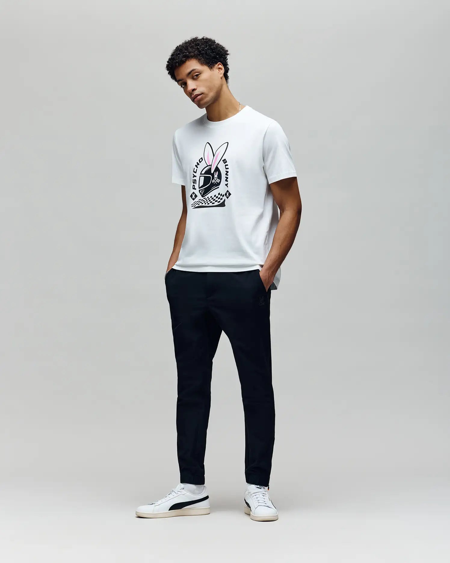 A man poses against a light gray background wearing the Psycho Bunny MENS RACING BUNNY GRAPHIC TEE (MT0200245), featuring a large bunny logo, paired with dark pants and white sneakers. His hands are in his pockets with a relaxed stance. (4)