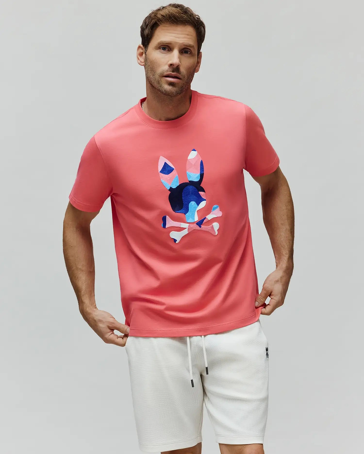 Mens Ara Graphic Tee in coral pink jersey knit, featuring a classic fit and a large central chest graphic of the iconic Psycho Bunny skull logo with blue, light blue, pink, and white geometric accents. (4)