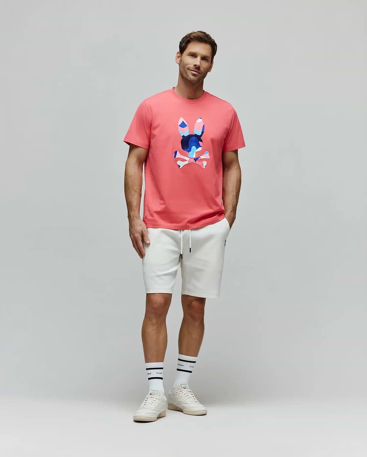 Mens Ara Graphic Tee in coral pink, a classic fit jersey with a large, center chest graphic bunny skull logo in blue, white, and pink abstract patterns. White relaxed fit french terry shorts. (5)