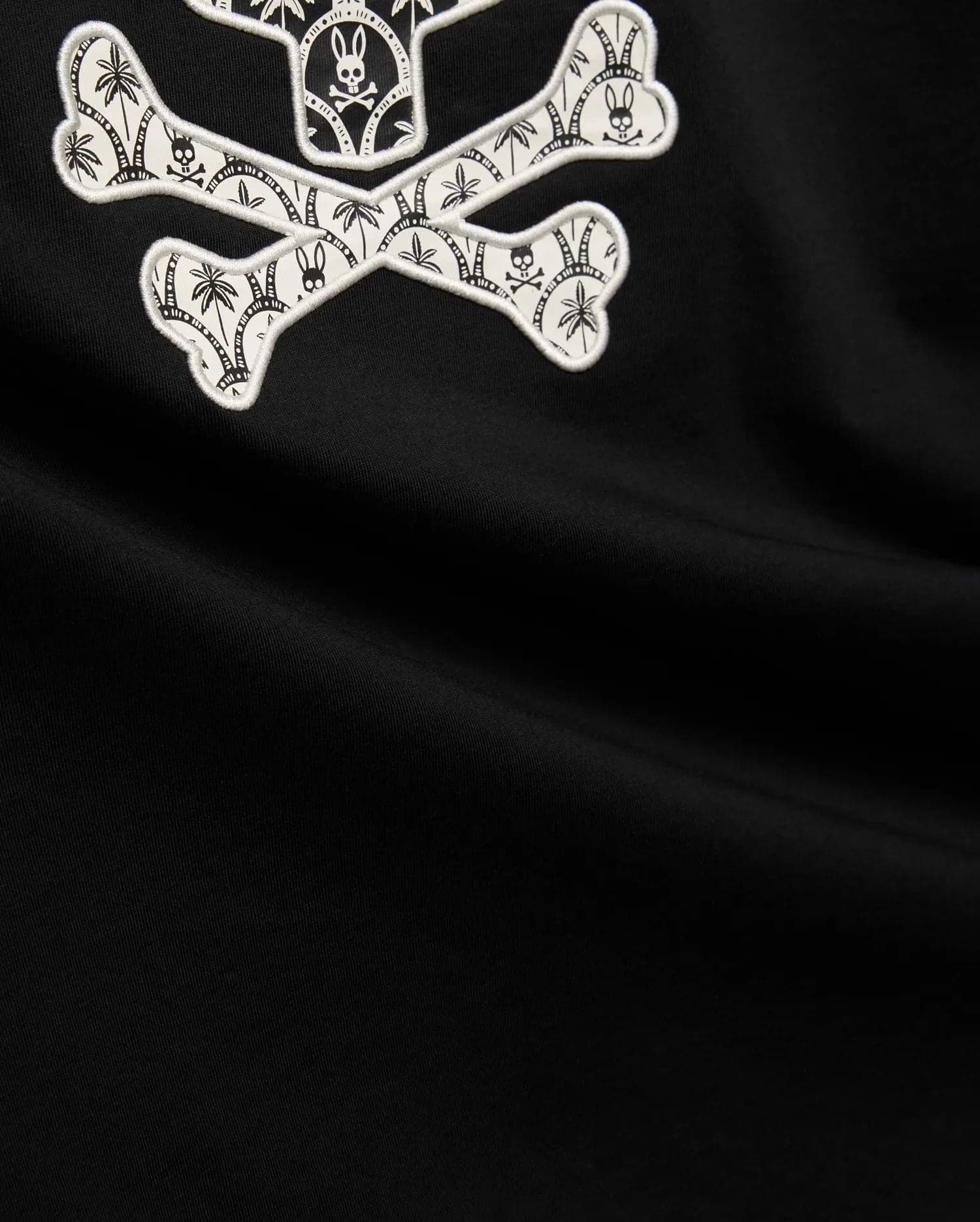 Mens Brad Graphic Tee, black smooth jersey with a large white embroidered Psycho Bunny skull and crossbones graphic, featuring tropical palm trees and mini bunny skulls within the bones. (6)