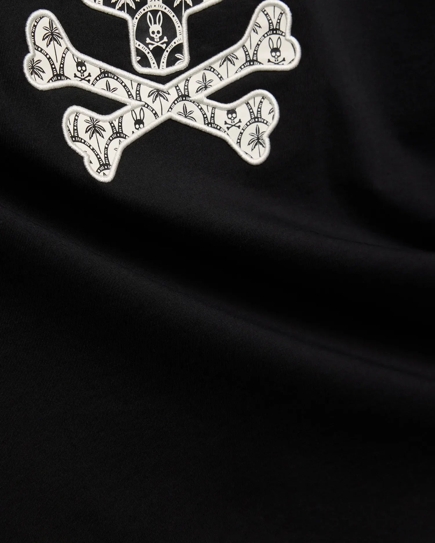 Mens Brad Graphic Tee, black smooth jersey with a large white embroidered Psycho Bunny skull and crossbones graphic, featuring tropical palm trees and mini bunny skulls within the bones. (6)