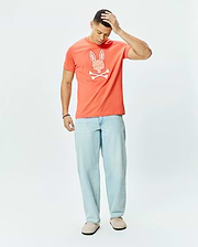 Mens Brad Graphic Tee, a coral classic fit jersey t-shirt featuring a large white patterned Psycho Bunny skull and crossbones graphic on the chest.