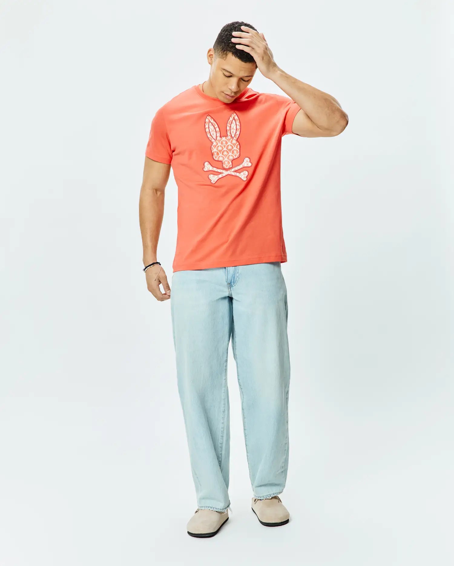 Mens Brad Graphic Tee, a coral classic fit jersey t-shirt featuring a large white patterned Psycho Bunny skull and crossbones graphic on the chest.