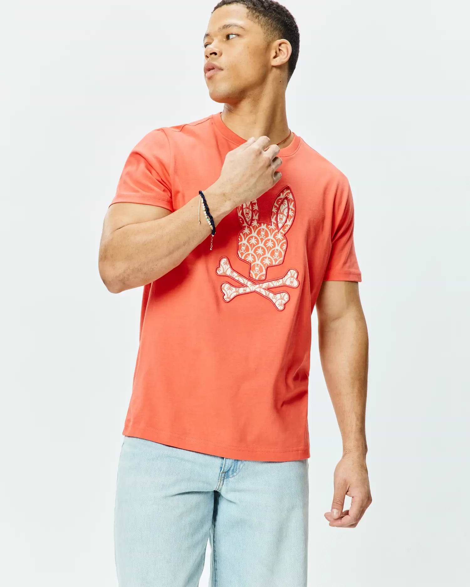 Mens Brad Graphic Tee, vibrant coral classic fit jersey t-shirt featuring a large central white Psycho Bunny skull and crossbones graphic with intricate patterned fill.
