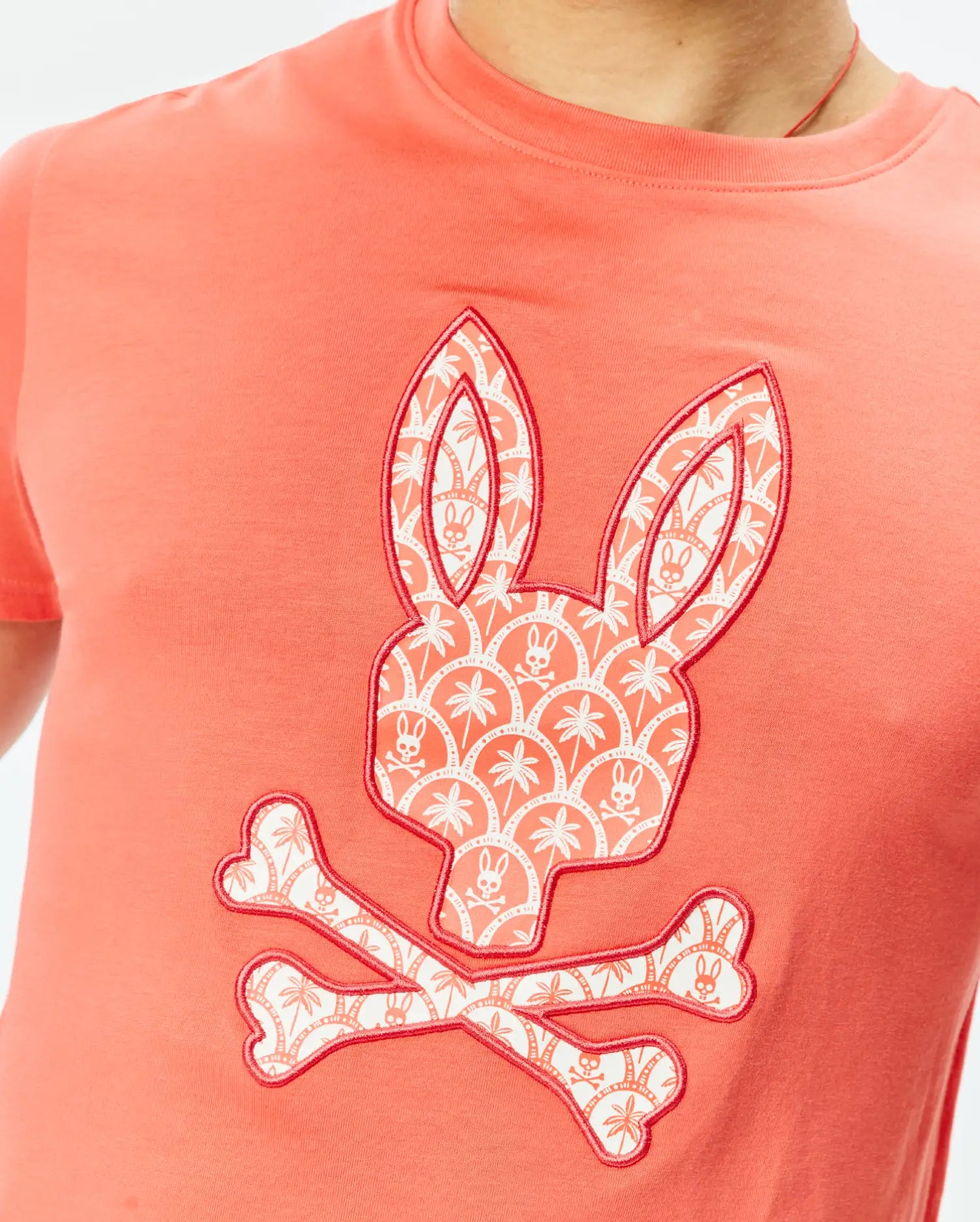 Mens Brad Graphic Tee, coral classic fit jersey t-shirt featuring a large central Psycho Bunny skull logo graphic, filled with a white palm tree and mini bunny skull pattern, outlined in red.