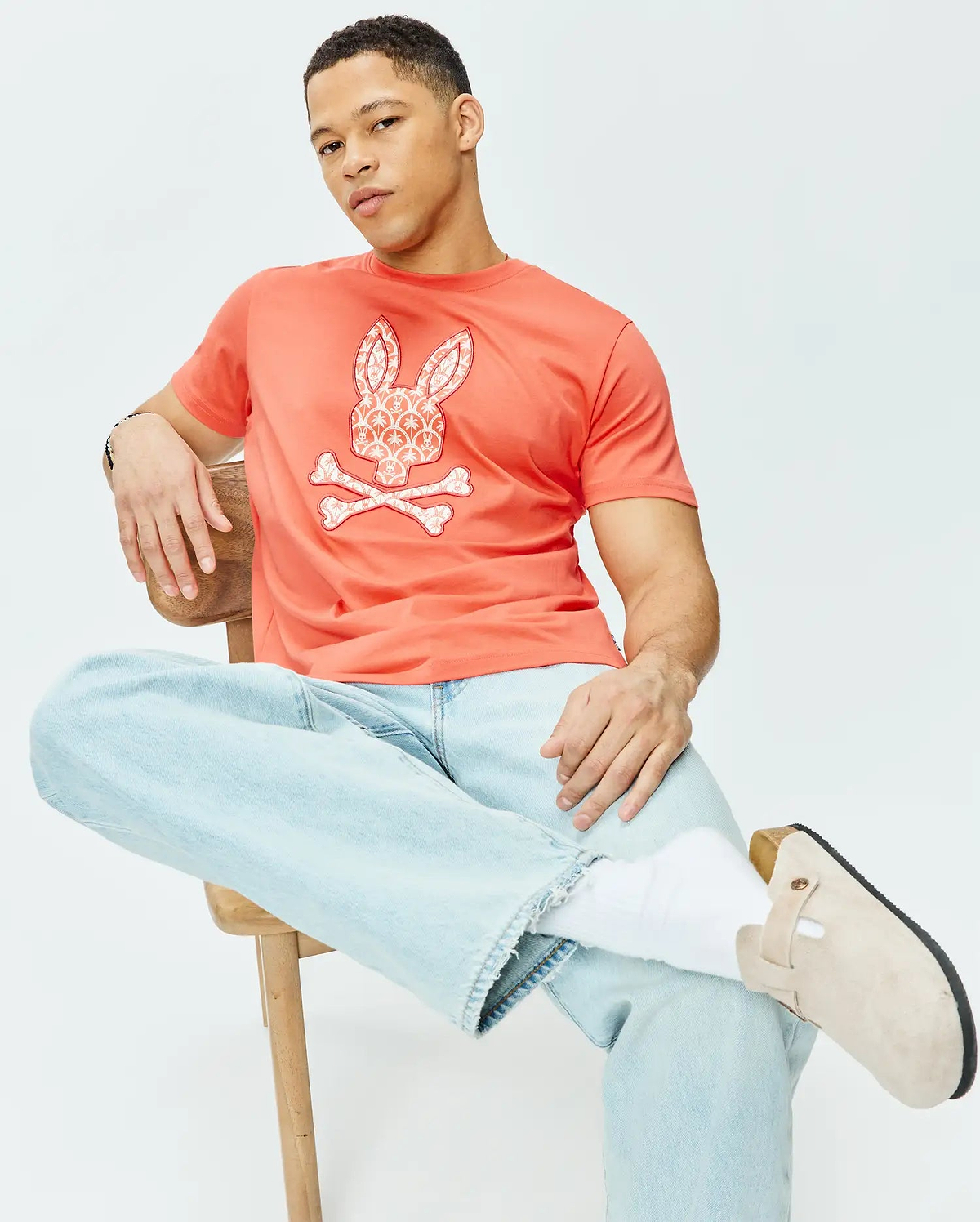 Mens Brad Graphic Tee in vibrant coral jersey knit, offering a classic fit and a prominent white Psycho Bunny skull and crossbones logo on the chest, detailed with an intricate palm tree pattern.