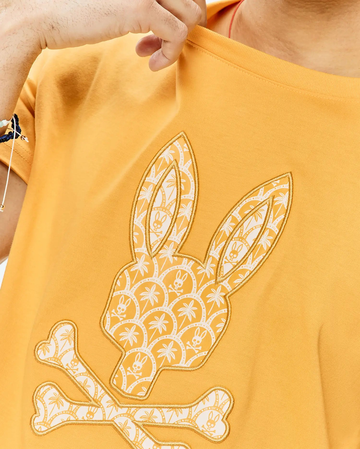 Mens Brad Graphic Tee. Mustard yellow classic fit jersey t-shirt with a large gold-outlined Psycho Bunny skull graphic on the chest, filled with white palm tree and bunny skull patterns.