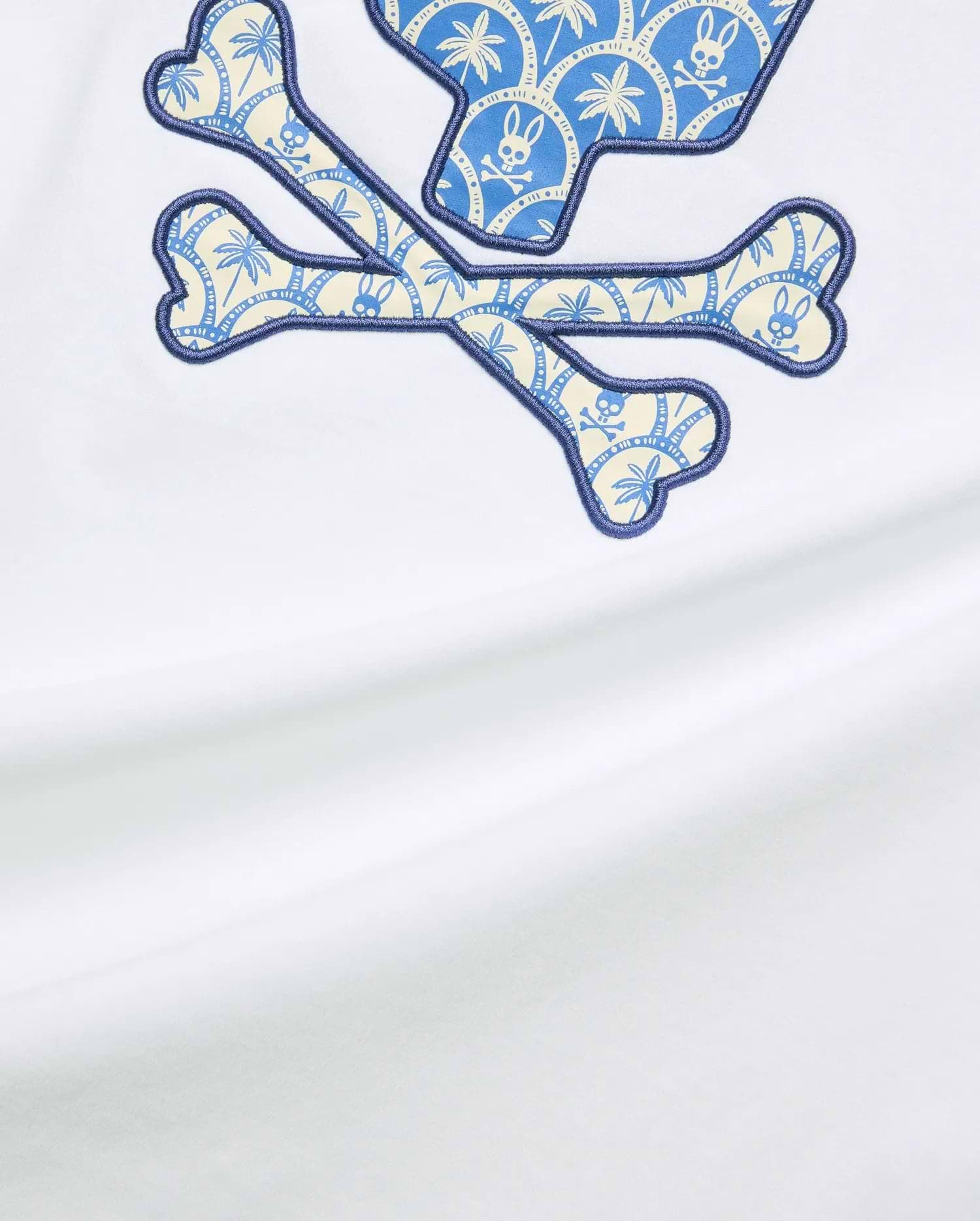 White Mens Brad Graphic Tee, classic fit, smooth jersey, featuring a large central Psycho Bunny skull and crossbones graphic with blue and cream palm tree and mini bunny skull pattern, outlined in dark blue embroidery. (5)