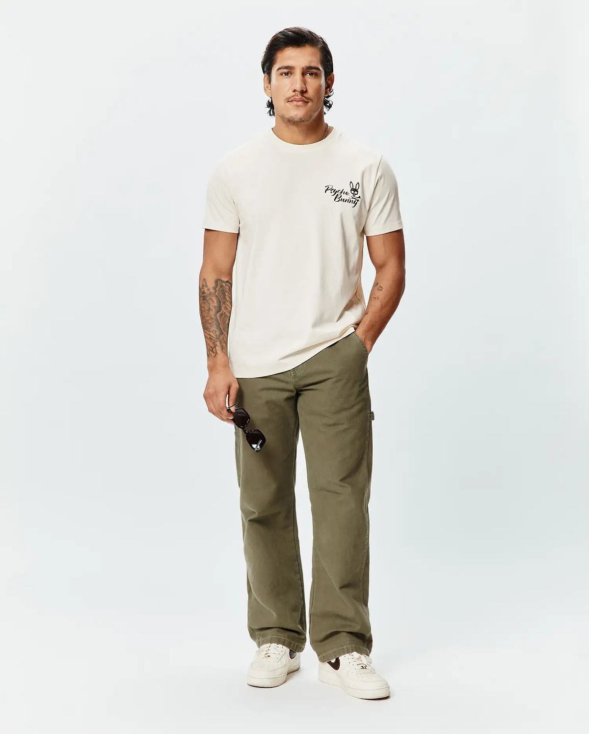 Mens Colin Back Graphic Tee, a classic fit ecru jersey t-shirt featuring a small, bold black Psycho Bunny skull logo and text printed on the left chest.