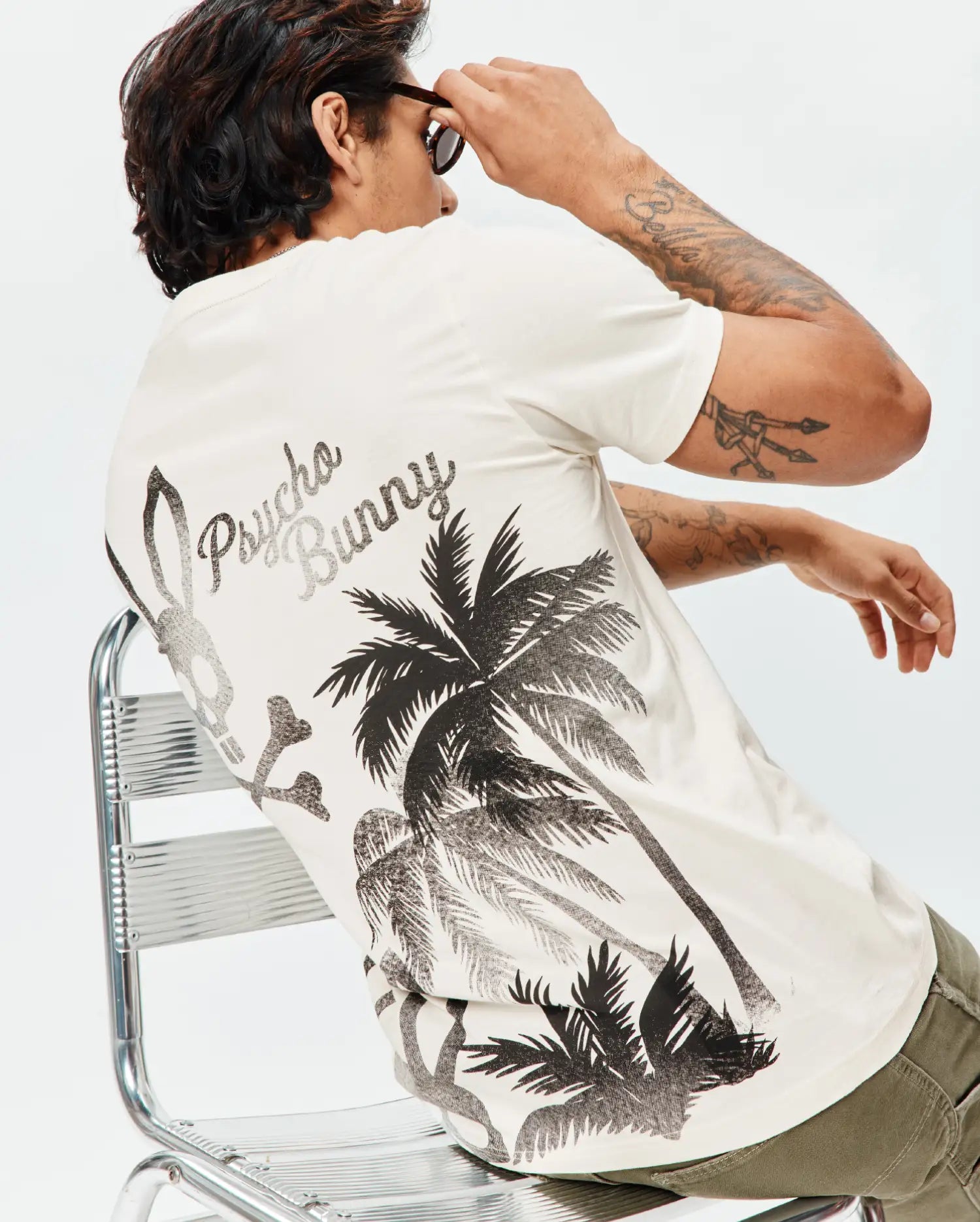 Mens Colin Back Graphic Tee, an ecru classic fit jersey knit t-shirt with a large black and grey back graphic featuring Psycho Bunny script, a bunny skull, palm trees, and crossbones.
