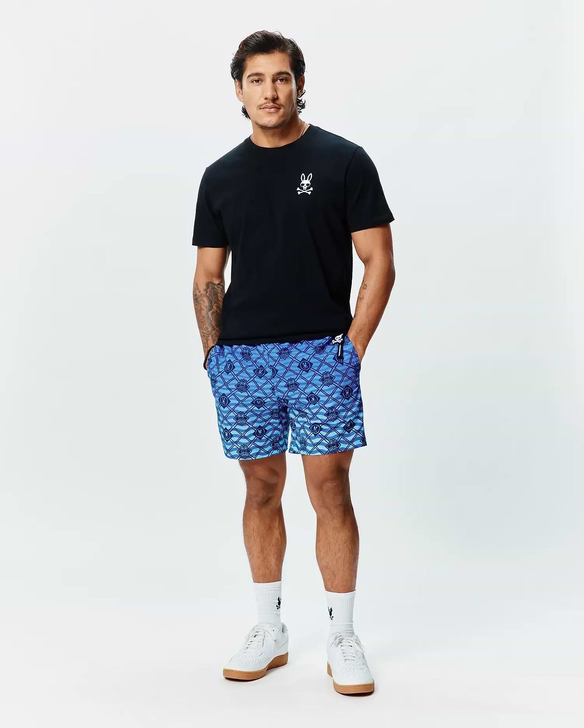 Mens Sunny Chest Graphic Tee, a classic fit black jersey t-shirt with a small white embroidered Psycho Bunny skull logo on the left chest, paired with blue geometric print shorts and white crew socks. (2)