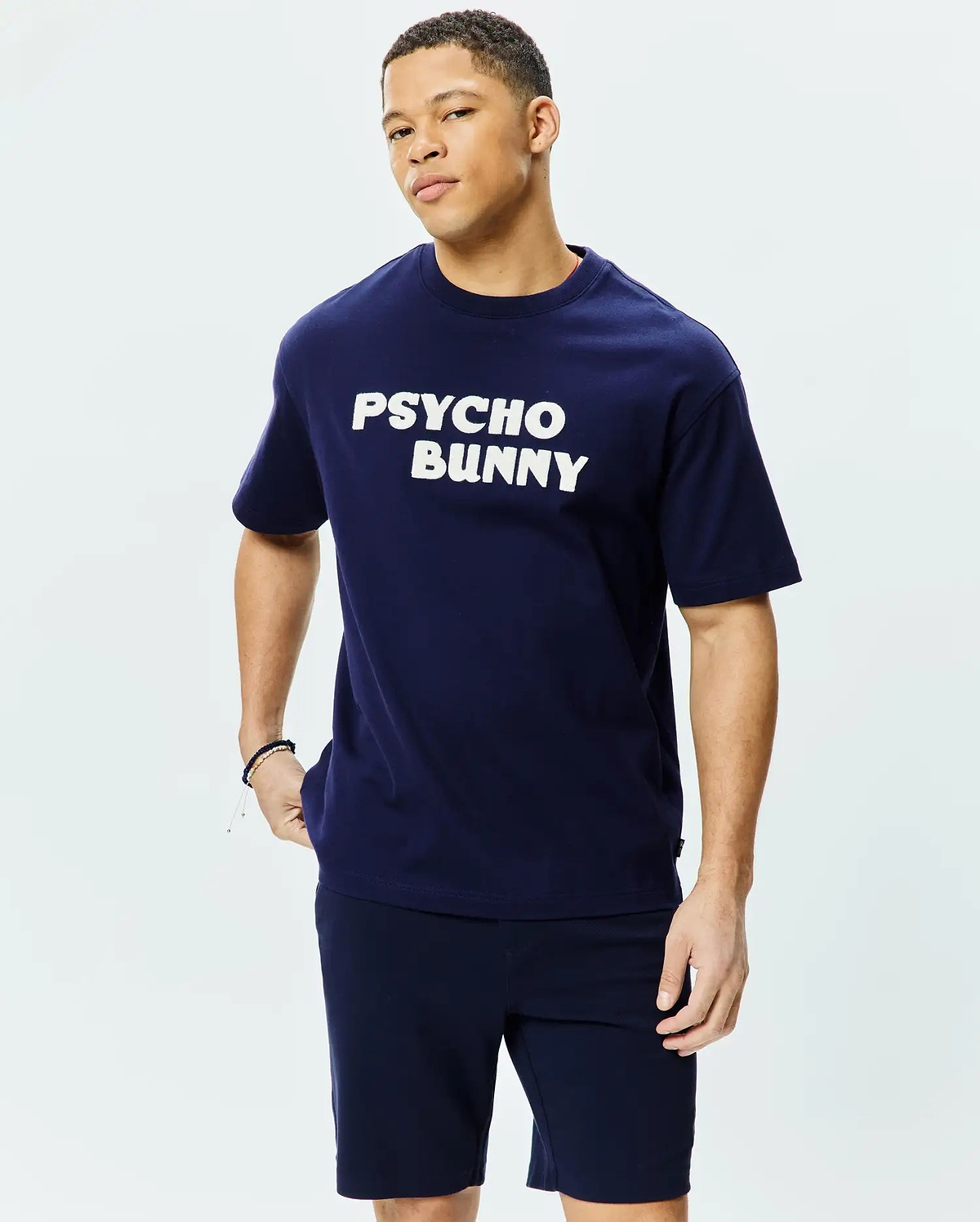 Mens Craig Wordmark Tee, a classic fit navy blue jersey t-shirt featuring a large white PSYCHO BUNNY wordmark graphic centered on the chest, with a subtle red inner collar trim and a small black brand tag on the left side seam. (1)
