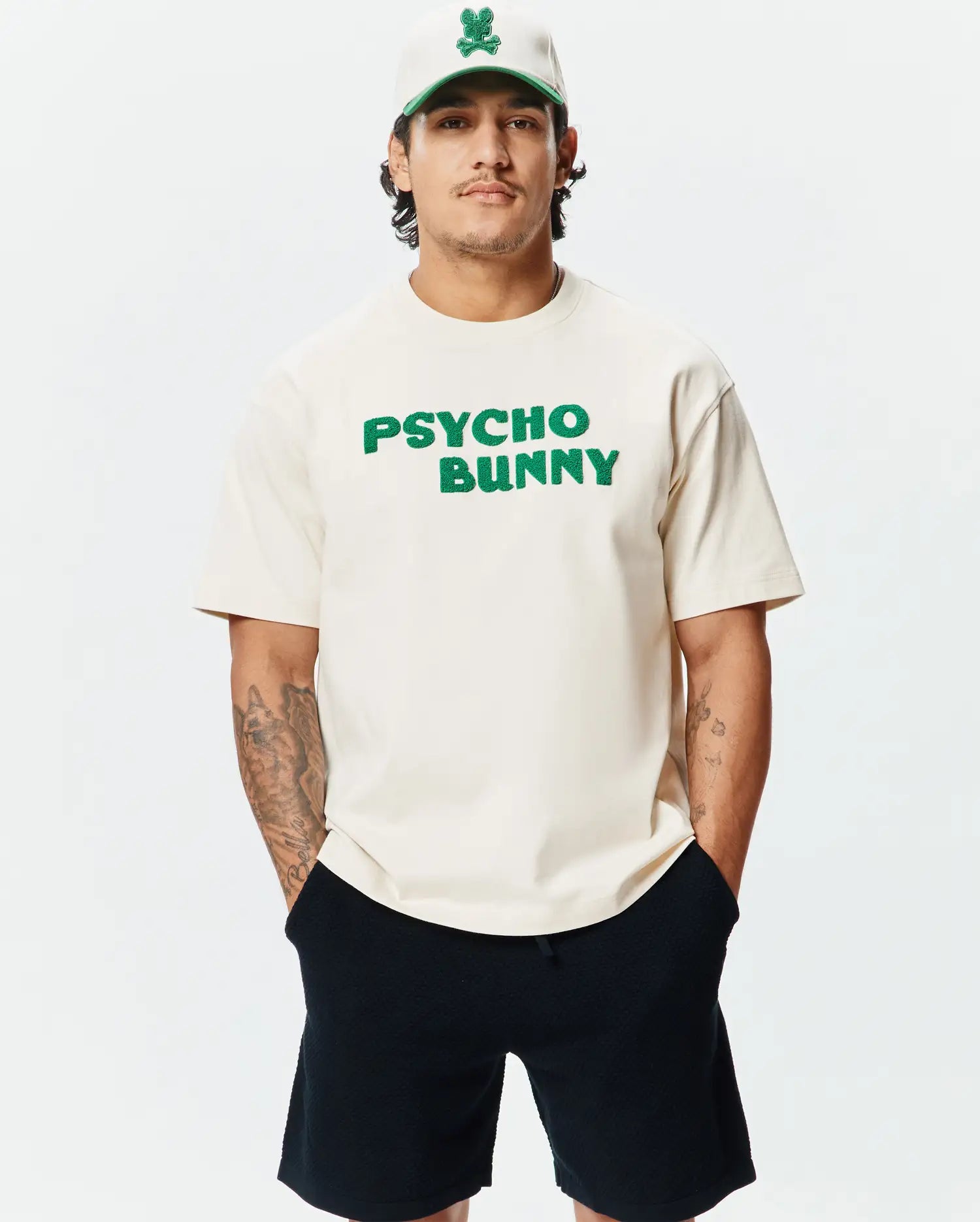 Mens Craig Wordmark Tee in cream jersey with a relaxed fit, featuring a large green textured PSYCHO BUNNY chest logo, complemented by a cream cap with a green brim and embroidered bunny skull.