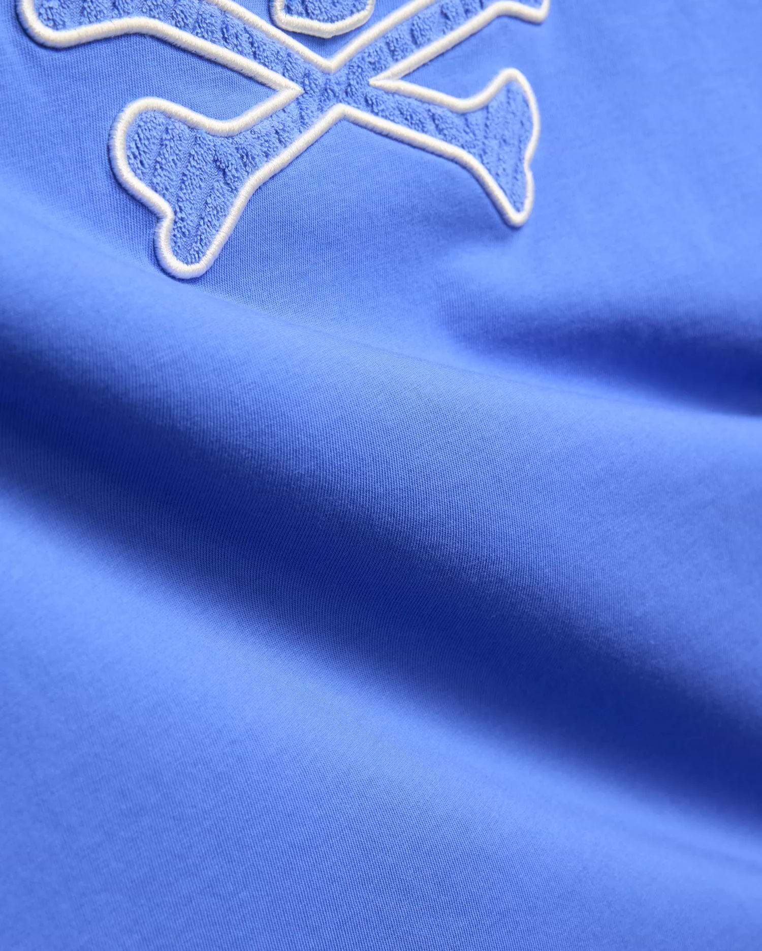 Mens Craig Graphic Tee in vibrant cornflower blue jersey knit, featuring a large, prominent white-outlined terry cloth embroidered crossbones logo. (6)