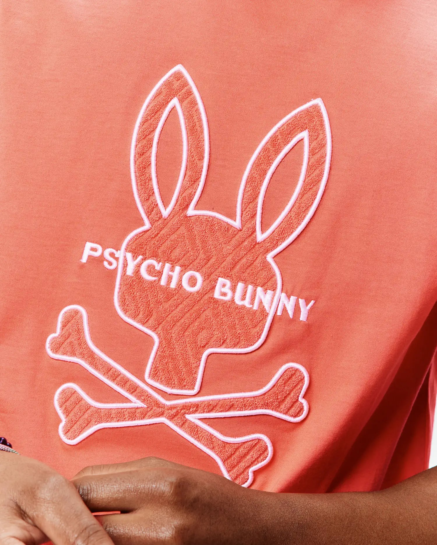 Mens Craig Graphic Tee, a coral jersey t-shirt with a large central white-outlined, textured coral Psycho Bunny skull and crossbones logo and white PSYCHO BUNNY text. (5)