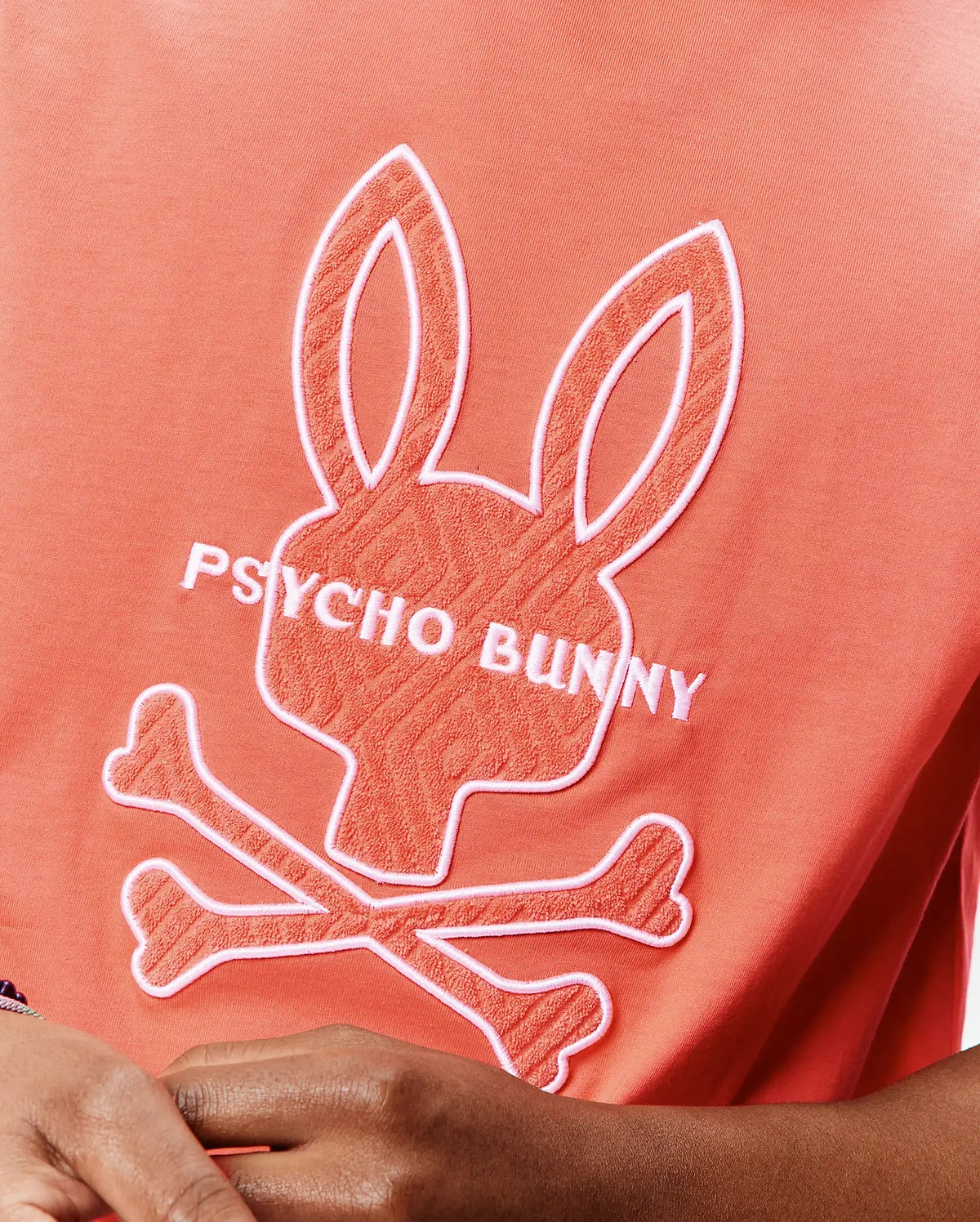 Mens Craig Graphic Tee, a coral jersey t-shirt with a large central white-outlined, textured coral Psycho Bunny skull and crossbones logo and white PSYCHO BUNNY text. (5)
