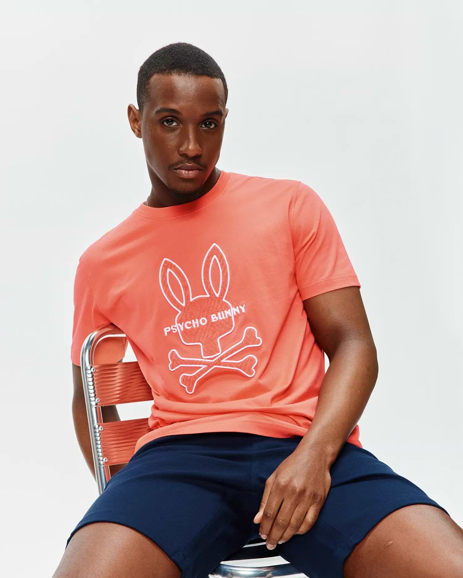 Mens Craig Graphic Tee, a coral classic fit jersey t-shirt featuring a large white Psycho Bunny skull and crossbones graphic with text on the chest. (3)