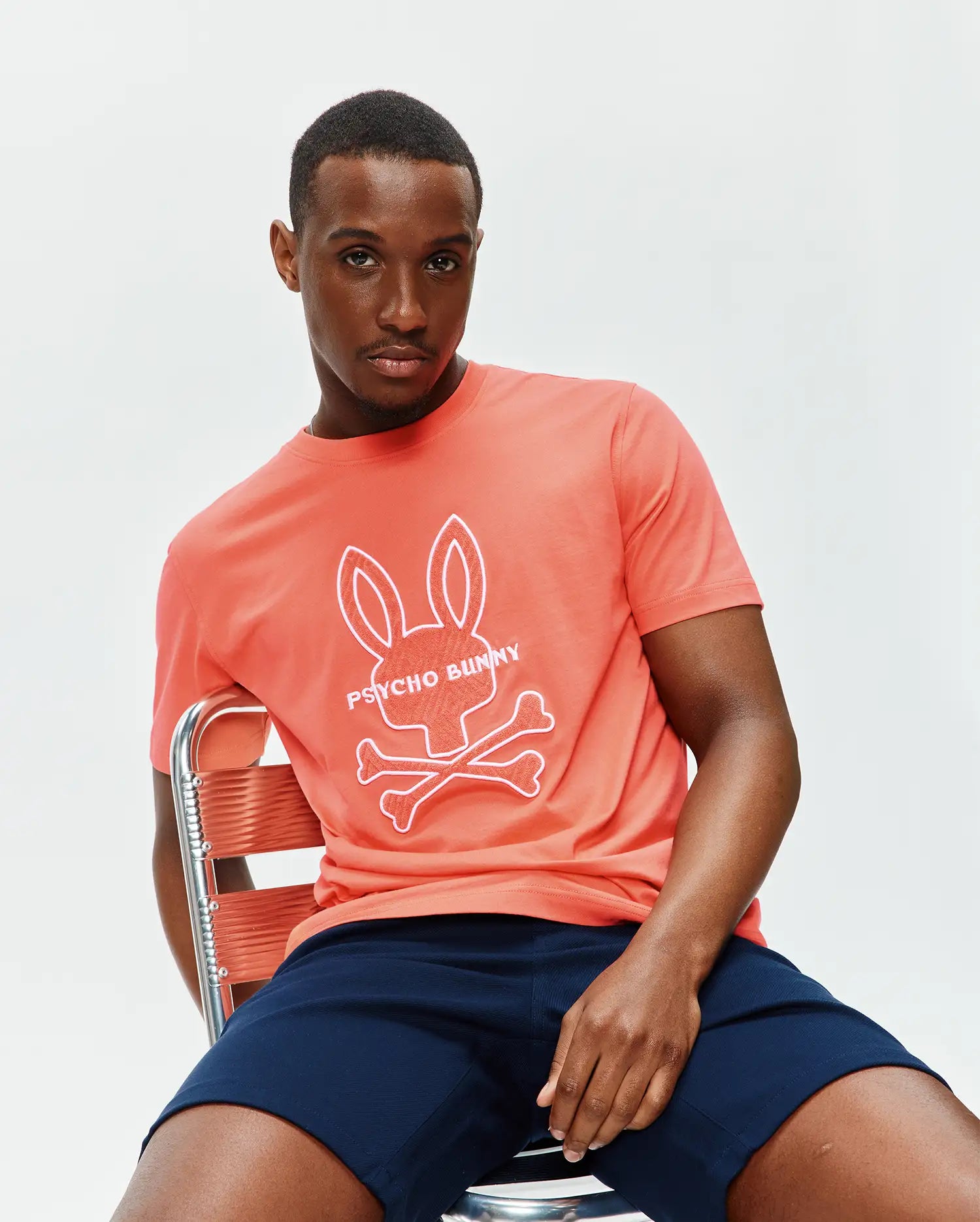 Mens Craig Graphic Tee, a coral classic fit jersey t-shirt featuring a large white Psycho Bunny skull and crossbones graphic with text on the chest. (3)